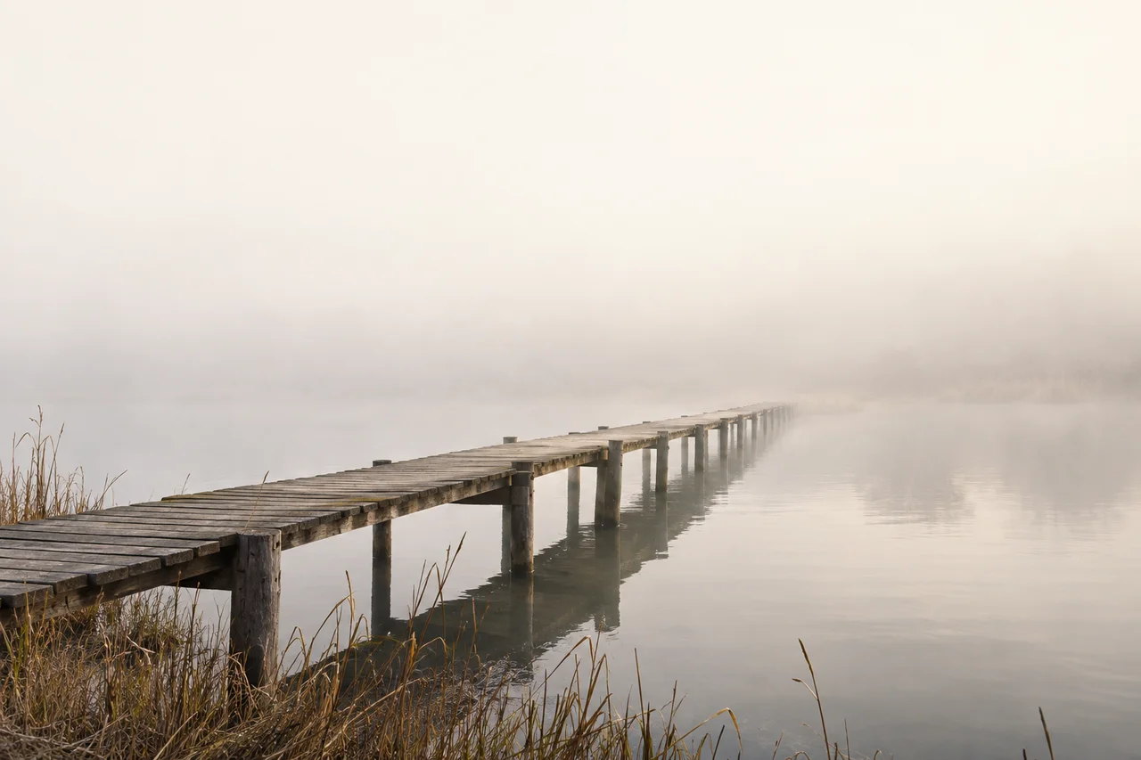 A timber footbridge extends across still water toward a distant shore in morning mist.